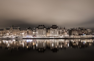 En nattvy över en stad vid vattnet, där upplysta fasader av stilfulla byggnader reflekteras i det stilla vattnet. Båtar ligger förtöjda längs kajen, och gatubelysningen skapar en varm och stämningsfull atmosfär under en molnig himmel. Bloggsida - Kristoffer Samsing